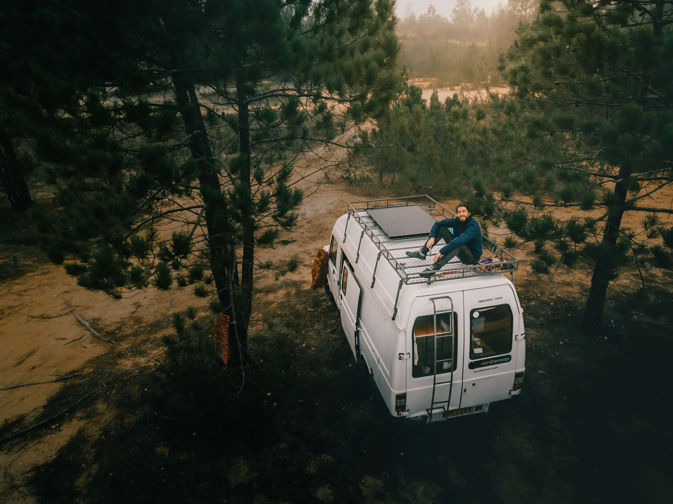 person sat on top of campervan roof, parked amongst shady pine trees